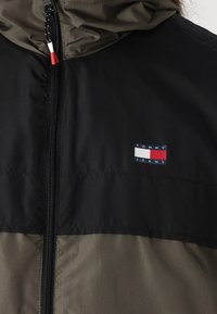 Juodas ir alyvuogių žalios vėjo nepraleidžiantis džemperis su užtrauktuku, reguliuojama gobtuvu ir mažu "Tommy Jeans" logotipo pleistru ant krūtinės.