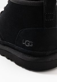 Svart mocka känga med texturerad överdel, rundad form, gummisula och präglad UGG-logga på hälen. Har kontrasterande sömmar.