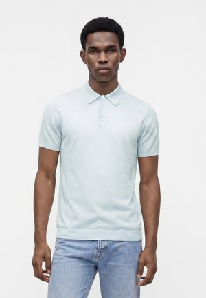 Ung mand med kort krøllet hår iført en lyseblå kortærmet polo og lyseblå jeans står foran en ensfarvet baggrund.