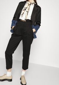 Svarta skräddarsydda byxor, vit skjorta, svart kavaj med denimärmar, krämfärgad scarf, beige skor och vita strumpor. Guldaccessoarer på bältet.