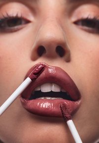 Płynna szminka o błyszczącym wykończeniu nałożona na pełne usta. Odcień to głęboki fioletowy róż, aplikator typu doe-foot w użyciu.