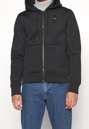 Mann trägt einen schwarzen Kapuzenpullover mit Reißverschluss, Fronttaschen, Kordelzug und kleinem gelben Logo auf der Brust, kombiniert mit blauen Jeans.