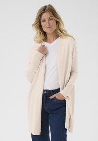 Cárdigan abierto de color beige claro hecho de un tejido de punto suave, con mangas largas y puños acanalados, llevado sobre una camiseta blanca de cuello en V y jeans azules.