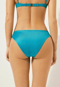 Bas de bikini turquoise en tissu lisse et léger, avec une coupe basse et des détails froncés sur les côtés.