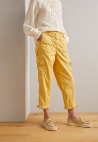 Persoon in gele cropped cargo broek, beige moccasin schoenen en een witte bloempatronen blouse met lange mouwen, staande op een houten vloer.