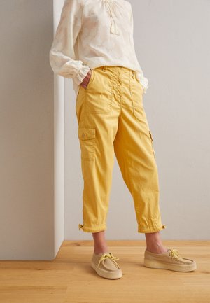 Persona che indossa pantaloni cargo gialli tagliati, mocassini beige e una blusa bianca a maniche lunghe con motivi floreali, in piedi su un pavimento di legno.