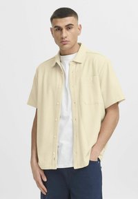 Beige overhemd met korte mouwen, knoopsluiting en een gestructureerde stof, met een enkele borstzak en een afgeronde zoom, gedragen over een wit T-shirt.