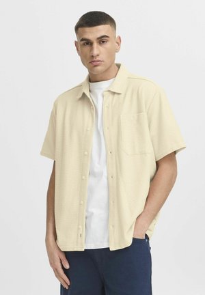 Chemise à manches courtes beige avec tissu texturé, poche poitrine unique et ourlet arrondi, portée par-dessus un T-shirt blanc.