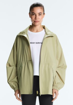 Žena oblečená v svetlozelenej nadrozmernej bunde na zips cez biele tričko s čiernym nápisom, stojaca s neutrálnym výrazom, jednoliate pozadie.