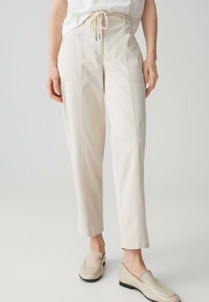 Persona che indossa pantaloni dritti a vita alta color avorio con coulisse e scarpe slip-on abbinate, in piedi davanti a uno sfondo neutro.