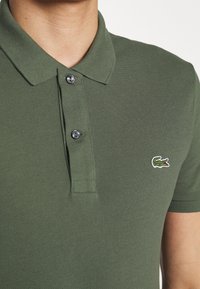 Polo vert en tissu texturé ; possède un col boutonné et un petit logo crocodile brodé en blanc et vert.