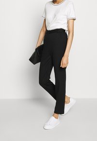 Mujer vestida con pantalones negros de talle alto, camiseta blanca con ribete negro, zapatillas blancas y sosteniendo un clutch negro.