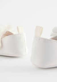 Weiße Babyschuhe mit weichem Stoffbezug, runder Form, dekorativen Blumenakzenten und kleinen Stoffschlaufen an der Rückseite für ein einfaches Anziehen.
