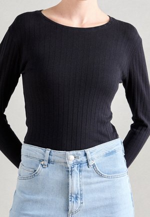 Femme portant un haut à manches longues côtelé noir rentré dans un jean taille haute bleu clair sur un fond neutre.