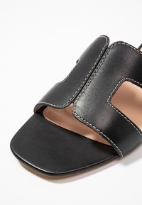 Black leather sandal featuring an open design with cutouts, contrasted stitching, a square toe, and a smooth texture.