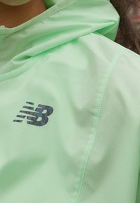 Jasnozielona kurtka wykonana z gładkiego, lekkiego materiału. Z przodu znajduje się czarne, wzorzyste logo New Balance. Projekt z kapturem.