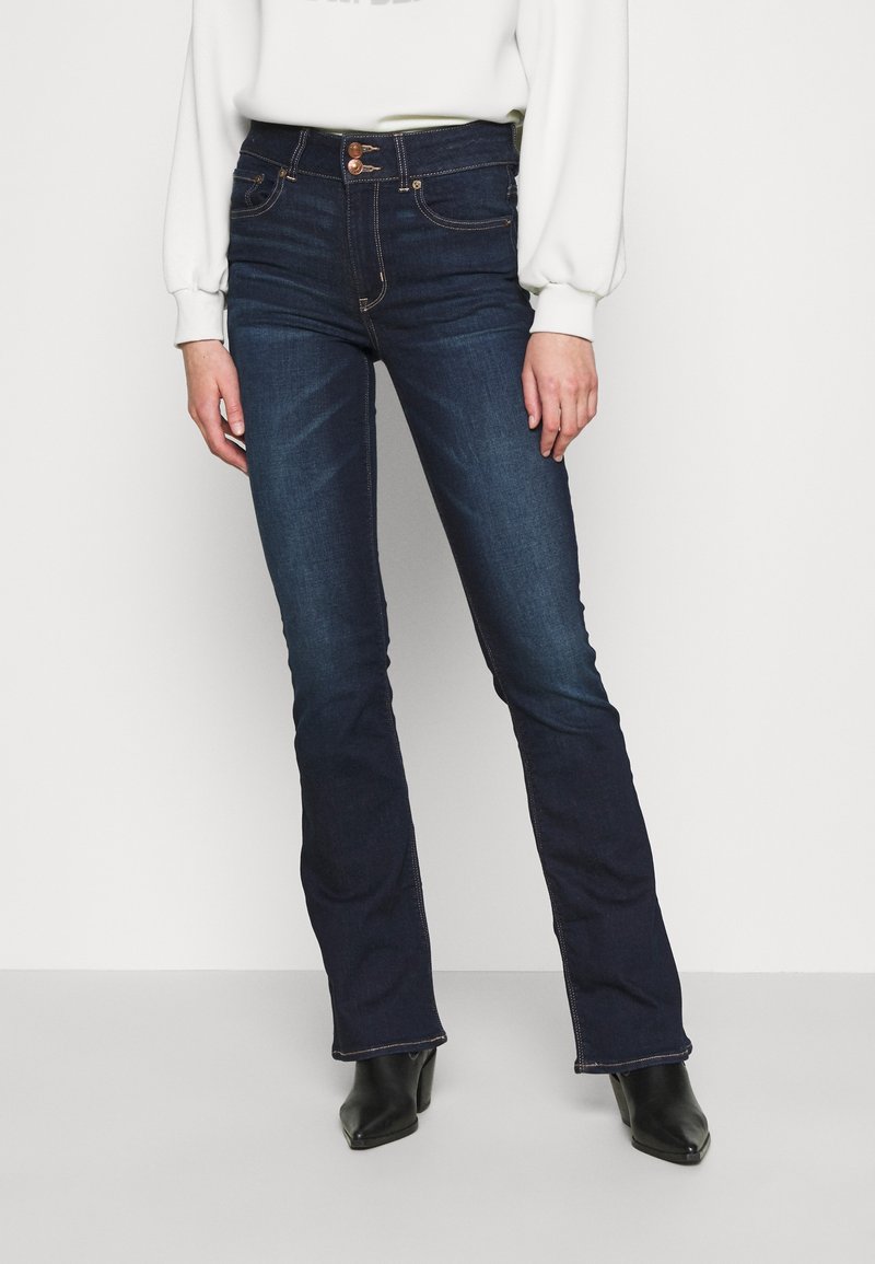 Jeans évasés bleu foncé en denim, avec une taille haute, des coutures claires et des poches avant, portés avec un sweat-shirt blanc et des bottes noires.