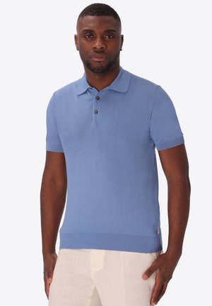 Uomo che indossa una polo azzurro chiaro a maniche corte e pantaloni beige, in piedi davanti a uno sfondo chiaro uniforme, leggermente rivolto in avanti.