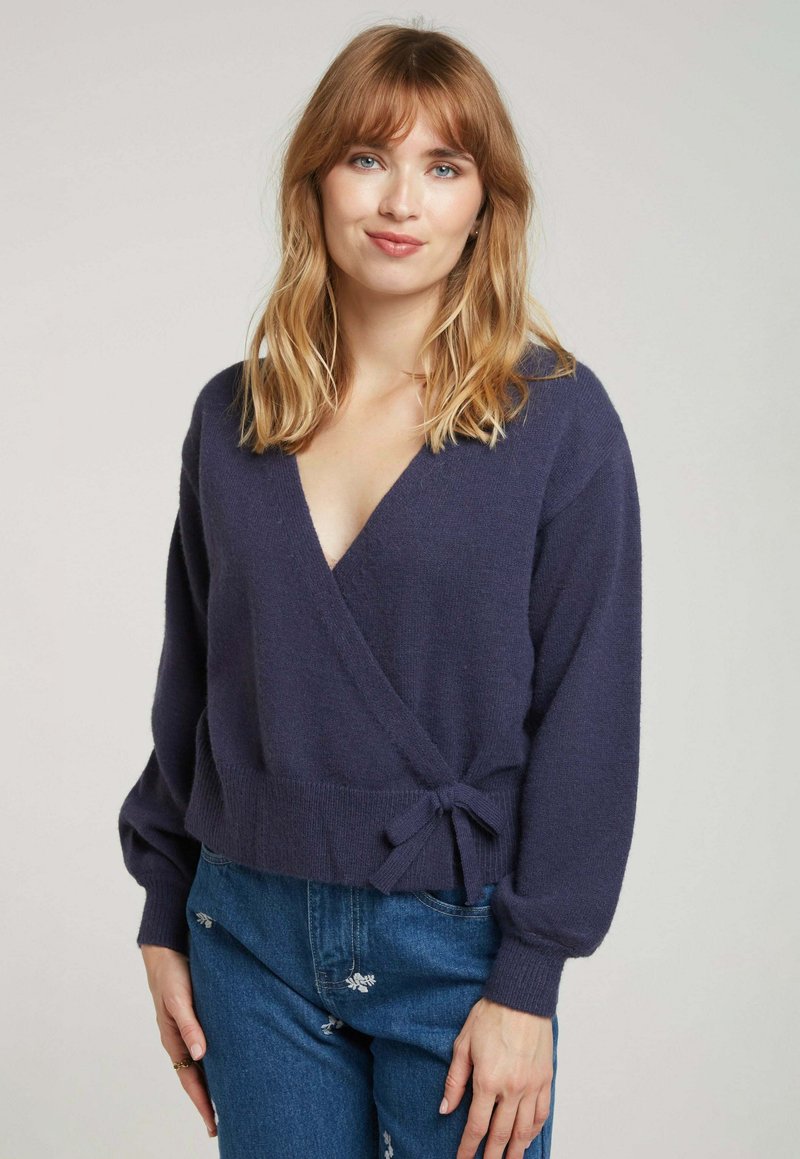 Cardigan navy en tissu tricoté doux, avec un décolleté en V profond, des manches ballon et une taille nouée. Assorti à un jean en denim bleu.