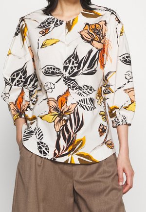 Blouse blanche avec grand imprimé floral orange, jaune et noir, manches trois-quarts bouffantes, associée à un pantalon marron ample.
