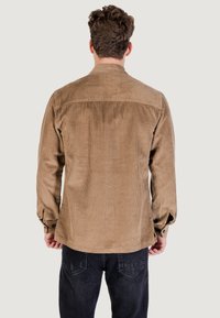 Camicia in corduroy marrone con righe verticali, polsini con bottone e orlo dritto, indossata sopra jeans scuri, vista da dietro.