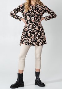 Tunique noire à motif floral avec manches longues, ornée de fleurs roses et crème. Associée à un pantalon crème ajusté et des bottines noires.