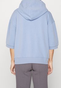 Osoba v světle modré mikině s krátkým rukávem a šedých žebrovaných kalhotách, zobrazená zezadu na bílém pozadí.