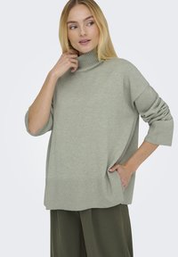 Suéter sobredimensionado verde claro con cuello de tortuga acanalado y puños. Textura suave, corte holgado, bolsillos laterales, combinado con pantalones verde oscuro.