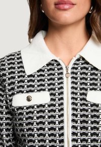 Giacca con motivo a fantasia in bianco e nero, con colletto bianco, chiusura con zip, due tasche frontali e bottoni rotondi argentati. Tessuto strutturato.