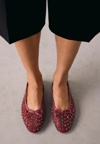 Ballerines en cuir rouge avec des œillets et un nœud à la pointe. Texture lisse, forme arrondie et design discret.