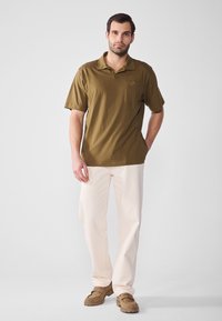 Olivgrön kortärmad polo med en ficka, kombinerad med ljuskrämfärgade byxor och beigea slipskor. Mjuk textur, avslappnad passform.