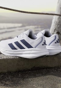 Chaussures de course blanches avec des rayures bleu foncé et des semelles rembourrées posées sur un rebord en pierre à l'extérieur près d'une corde et d'un tronc d'arbre.