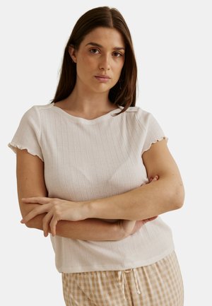 Femme aux longs cheveux bruns portant un haut blanc côtelé à manches courtes et un pantalon beige à carreaux vichy, bras croisés, expression neutre, fond uni.