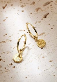 Orecchini a cerchio in oro con un ciondolo circolare caratterizzato da un design di occhio inciso. Il ciondolo ha linee radiali e pende dal cerchio.