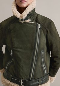 Groene suede jas met een schapenvachtkraag, asymmetrische ritssluiting, ritszakken aan de zijkant en een ceintuur om de taille. Textuur en aansluitend ontwerp.