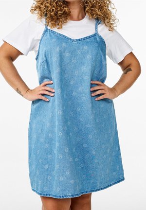 Kobieta ubrana w jasnoniebieską, bezrękawnikową sukienkę jeansową z białymi kwiatowymi wzorami na białej krótkim rękawie, ręce na biodrach, widoczne kręcone włosy.