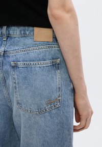 Jean en denim bleu clair avec un patch en cuir à la ceinture et des coutures dorées sur la poche arrière ornée d'un logo de la marque.