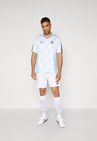  Lichtblauw geruite voetbalshirt met zwarte accenten, witte shorts en witte sokken. Heeft een zwart en blauwe kraag, logo-details en een gestroomlijnde pasvorm.