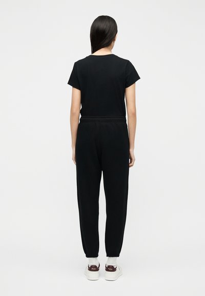 Γυναίκα με μακριά μαύρα μαλλιά, που φορεί μαύρο t-shirt, μαύρο παντελόνι jogging, λευκές κάλτσες και λευκά αθλητικά παπούτσια με καφέ λεπτομέρειες, που κοιτάζει προς τα πίσω.