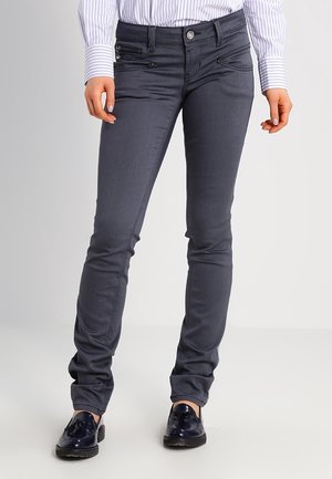 Tiimalasiharmaat slim-fit-farkut vetoketjulla varustetulla etutaskulla, yhdistetty raitapaitaan ja tummansinisiin hapsuja sisältäviin loafer-kenkiin, seisovalla henkilöllä.