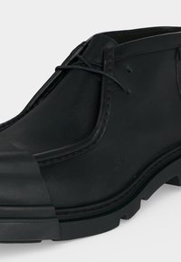 Bottine noire en cuir avec bout arrondi, coutures minimales, lacets fins et semelle épaisse en caoutchouc avec motif de bande de roulement.