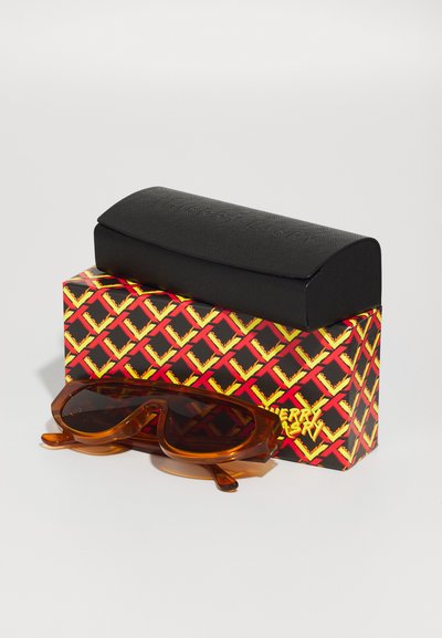 Amber sunglasses with a rounded design and dark lenses, placed on a patterned box featuring red, yellow, and black geometric designs.