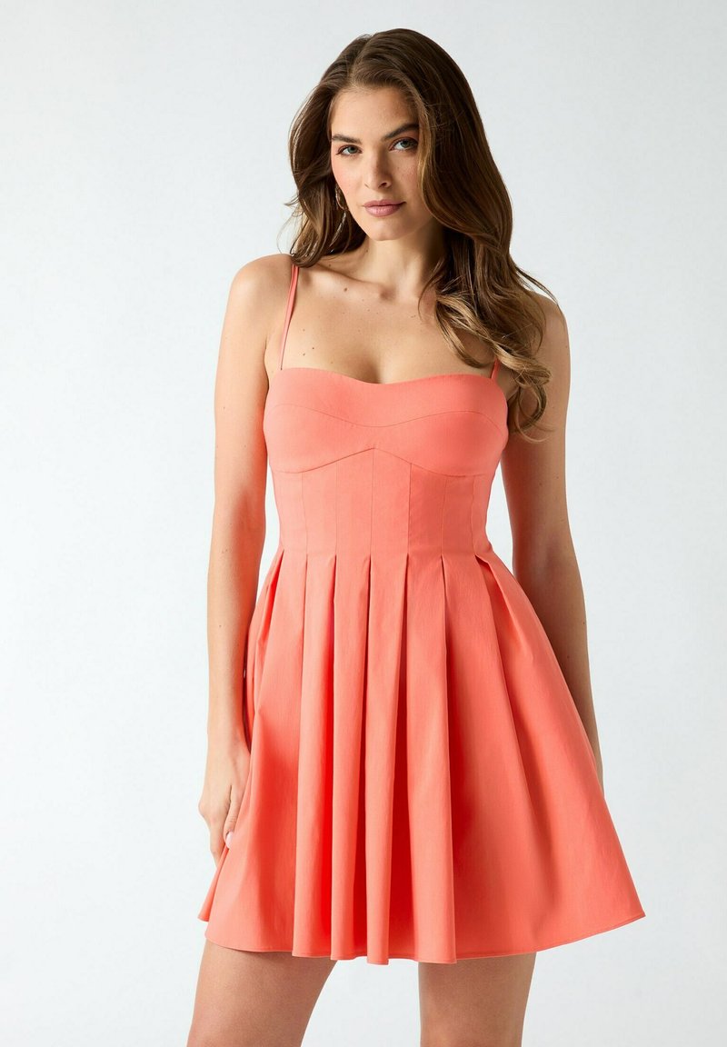 Femme aux longs cheveux bruns portant une robe corail plissée sans manches, debout sur un fond blanc uni.