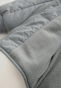 Weiche graue Jacke mit einer Kombination aus glattem Außenstoff und strukturiertem Fleece, sichtbaren Nähten und Reißverschlusselementen.