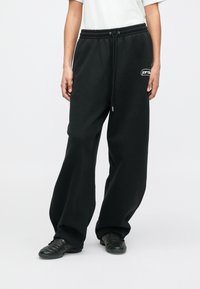 Άτομο φορώντας μαύρες φαρδιές φόρμες με κορδόνι στη μέση και μαύρα αθλητικά παπούτσια, που στέκεται μπροστά σε λευκό φόντο.