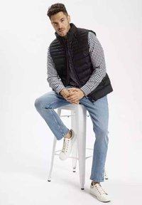 Czarna pikowana kamizelka nałożona na niebieską i białą koszulę w kratę, jasnoniebieskie dżinsy i białe sneakersy, siedząca na białym stołku barowym.