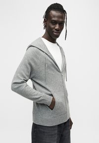 Sweat à capuche gris en tissu doux, avec fermeture éclair devant, poches kangourou et capuche à cordon. Porté sur une chemise blanche.