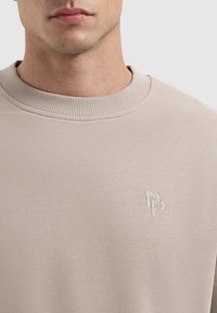 Beige sweatshirt met geribbelde ronde hals, voorzien van een gebosseld logo op de borst. De stof lijkt glad met een subtiele textuur.