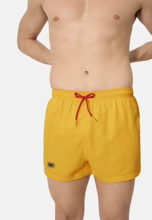 Hombre con pantalones de baño amarillos con cordón rojo, de pie con las manos detrás de la espalda, mostrado desde el pecho hasta la mitad del muslo.