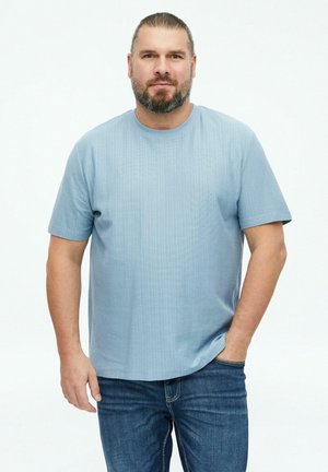 Man met baard en naar achter gebonden haar, draagt een lichtblauw shirt met korte mouwen en een blauwe spijkerbroek, staand met één hand in de zak op een witte achtergrond.